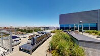 This rooftop amenity space combines a well-appointed interior lounge with a generous outdoor terrace, offering a dynamic environment for relaxation and socializing. The indoor area features a contemporary open-plan design with light hardwood flooring, recessed lighting, and large floor-to-ceiling windows that frame panoramic city views and flood the space with natural light. A bold cityscape mural wraps across the walls, adding a vibrant urban aesthetic. The interior includes a spacious lounge seating area with plush sectional sofas, accent pillows, and a wall-mounted television framed by warm wood paneling. A central white island with modern barstools serves as a gathering point, complemented by a sleek wet bar and a dramatic rod pendant light fixture overhead. Additional seating nooks positioned beside corner windows provide quieter vantage points overlooking the green roof. The outdoor terrace extends the living space significantly, featuring a green roof planted with ornamental grasses and native plantings that create a naturalistic atmosphere. Comfortable lounge seating with striped cushions, low coffee tables, a fire bowl planter, and a built-in bar counter are thoughtfully arranged to maximize the sweeping views of the surrounding cityscape and open sky.