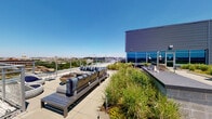 This rooftop amenity space combines a well-appointed interior lounge with a generous outdoor terrace, offering a dynamic environment for relaxation and socializing. The indoor area features a contemporary open-plan design with light hardwood flooring, recessed lighting, and large floor-to-ceiling windows that frame panoramic city views and flood the space with natural light. A bold cityscape mural wraps across the walls, adding a vibrant urban aesthetic. The interior includes a spacious lounge seating area with plush sectional sofas, accent pillows, and a wall-mounted television framed by warm wood paneling. A central white island with modern barstools serves as a gathering point, complemented by a sleek wet bar and a dramatic rod pendant light fixture overhead. Additional seating nooks positioned beside corner windows provide quieter vantage points overlooking the green roof. The outdoor terrace extends the living space significantly, featuring a green roof planted with ornamental grasses and native plantings that create a naturalistic atmosphere. Comfortable lounge seating with striped cushions, low coffee tables, a fire bowl planter, and a built-in bar counter are thoughtfully arranged to maximize the sweeping views of the surrounding cityscape and open sky.