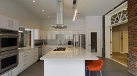 This single-floor space showcases a sleek, modern kitchen designed with both style and functionality in mind. The centerpiece is a generously sized island topped with striking white marble, complete with natural veining that adds visual depth. A gas cooktop is integrated directly into the island surface, complemented by a stainless steel island-mounted range hood that draws the eye upward toward the notably high ceilings, reinforcing the open, airy atmosphere.

The surrounding cabinetry is finished in crisp white with minimalist hardware, creating a clean, cohesive look. A shimmering tile backsplash adds subtle texture against the white surfaces, while stainless steel appliances, including a French door refrigerator and dishwasher, provide a polished, contemporary finish. Recessed lighting and a pendant light fixture illuminate the space effectively.

An exposed brick wall on one side introduces a warm, industrial contrast to the otherwise modern palette. Dark flooring grounds the space visually, while a bold orange accent chair injects a pop of color. The open-concept layout and elevated ceilings combine to create a spacious, inviting environment well-suited for both cooking and entertaining.