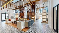 Located at 4401 Barclay Downs Dr in Charlotte, NC, this sophisticated amenity space makes an immediate impression with its soaring double-height ceilings and an abundance of natural light streaming through expansive floor-to-ceiling glass windows and industrial-style garage doors. The signature black-and-white patterned encaustic tile flooring flows seamlessly throughout, anchoring the space with a bold yet timeless aesthetic.

The main lounge area is defined by cascading crystal pendant chandeliers that add a luminous, artistic focal point overhead. Rich warm-toned wood slatted partitions divide the space elegantly, creating intimate seating areas furnished with curated lounge chairs, sofas, and accent tables. A standout linear fireplace set within a wood-paneled wall serves as a cozy gathering point, topped by a large-format display screen and flanked by sculptural décor pieces.

The fully equipped communal kitchen features a substantial, light-colored stone island with bar seating, a professional-grade range with a custom hood, and ample counter space — all bathed in natural light from the oversized exterior windows. A hallway adorned with framed artwork and the same signature tile flooring connects ancillary rooms, including restrooms and additional multi-use spaces. Together, these elements create a refined, welcoming, and design-forward environment.