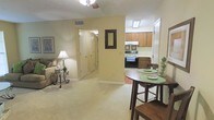 This single-floor residence is designed with an open-concept layout that seamlessly connects the living area, dining space, and kitchen, creating a cohesive and functional flow throughout the main living areas. The living room features soft, neutral carpet flooring, warm beige walls, and a ceiling fan with a light fixture overhead, contributing to a comfortable and well-lit environment. A decorative wall mirror with an ornate dark frame serves as a focal point on the shared wall between the living and kitchen areas. The dining area accommodates a round wooden bistro-style table with matching chairs, positioned conveniently at the edge of the open-plan space. The kitchen is equipped with wood-toned upper and lower cabinetry, a white electric range with an overhead range hood, and tile flooring that distinguishes it from the carpeted living areas. A hallway visible from the main living space leads to additional rooms, suggesting a practical layout with private spaces separated from the communal areas. The overall ambiance is warm and approachable, with consistent neutral tones throughout.