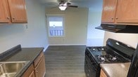This single-floor residence offers a functional and comfortable layout designed for everyday living. The kitchen is equipped with a black gas range and oven, dark granite-style countertops, and warm wood cabinetry, providing ample storage and workspace for cooking. A range hood overhead adds a practical touch to the culinary space. The kitchen flows seamlessly into an adjacent dining or living area, creating an open and connected feel throughout the main living space. This area is finished with durable hardwood-style laminate flooring in a medium gray-brown tone, lending a modern, cohesive aesthetic underfoot. A ceiling fan with an integrated light fixture is centrally mounted overhead, ensuring year-round comfort and efficient air circulation. Natural light filters into the space through a window framed in white trim, brightening the room and providing a view to the exterior. The walls are painted in a soft, neutral palette — a muted green-gray tone — that complements the flooring and cabinetry. White baseboards run along the perimeter, adding a clean, finished look. Overall, the space presents a straightforward, move-in-ready layout with practical features suited for comfortable single-floor living.