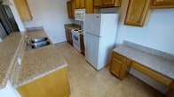 This single-floor space offers a practical and well-appointed layout designed for comfortable everyday living. The kitchen serves as a central feature, showcasing warm honey-toned wood cabinetry that provides generous storage both above and below the countertops. The counters themselves are finished in a light granite-style surface, offering ample workspace for meal preparation. White appliances — including a coil-top electric range, an over-the-range microwave, and a full-sized top-freezer refrigerator — are neatly integrated into the layout, contributing to a clean and cohesive look. A double sink is conveniently positioned along the perimeter, adding further functionality to the space. The kitchen flooring features neutral-toned tile in a subtle grid pattern, which is easy to maintain and complements the overall color palette. The walls are painted in a crisp, light neutral tone, keeping the space feeling bright and open. The layout allows for ease of movement throughout the kitchen, with an island or peninsula providing additional counter space and a natural dividing point between the kitchen and the rest of the living area. Overall, the space presents a tidy, functional environment suited to a variety of lifestyles.