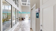 Located at 7000 University Blvd in Winter Park, Florida, this single-floor space offers a well-appointed layout comprising a living room and a dedicated office area. The living room serves as the centerpiece of the space, boasting impressively high ceilings with an elegant coffered detail and recessed lighting that draw the eye upward. Oversized windows flood the room with abundant natural light while offering views of the surrounding outdoor environment. The interior design features a cheerful coastal-inspired color scheme, with signature teal accent walls and trim complementing the neutral grey and white tones throughout. Large-format stone tile flooring flows seamlessly across the entire space, lending a clean and polished aesthetic. The living area is furnished with comfortable seating arrangements, including a sofa, accent chairs, and a central coffee table, anchored by a wall-mounted flat-screen television set against a warm wood-paneled accent wall. The office area provides a practical workspace equipped with a substantial wood desk, yellow upholstered chairs, and desktop computing, accented by colorful contemporary artwork above. Restroom facilities are also accessible within the space. The overall ambiance is bright, open, and inviting, with a modern yet comfortable feel throughout.