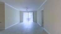 This single-floor residence features a clean, open layout designed to maximize both comfort and functionality. The main living area is finished with large-format ceramic tile flooring in a neutral tone, lending a polished, low-maintenance quality to the space. Crown molding lines the perimeter of the ceiling, adding a refined architectural detail to the otherwise understated design. A ceiling fan is centrally mounted, providing both air circulation and subtle visual interest overhead. Natural light fills the room through a full-width sliding glass door dressed with vertical blinds, offering convenient light control while connecting the interior to an outdoor area beyond. The smooth, light-toned walls contribute to an airy, open atmosphere throughout. A doorway visible to the right side of the living area leads to additional rooms, suggesting a practical and straightforward floor plan. The overall ambiance is bright and neutral, providing a versatile canvas well-suited for a variety of interior styles. The combination of tile flooring, crown molding, and abundant natural light gives the space a comfortable, inviting character consistent with warm-climate residential design.