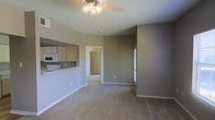 This single-story space features an open-concept layout that connects the living area to the kitchen through a pass-through window, creating a functional and visually connected floor plan. The living area is finished with wall-to-wall carpet in a neutral beige tone, complemented by taupe-painted walls and white baseboards that lend a clean, cohesive look throughout. A ceiling fan with a light fixture serves as the central overhead lighting source in the main living space, which benefits from natural light via a window along the far wall. The kitchen is accessible via the pass-through cutout and features light wood cabinetry, white appliances including a microwave and range, and a countertop that doubles as a breakfast bar. A hallway visible from the main living area leads to additional rooms, including what appears to be a bedroom with a white refrigerator visible just inside the doorway—suggesting a versatile layout. The textured ceiling adds a subtle architectural detail consistent throughout the space. Overall, the unit presents a straightforward, functional design with a neutral palette suited for a variety of furnishing styles.