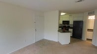This single-floor residential unit offers a clean, functional layout designed for comfortable everyday living. The open-concept design seamlessly connects the living area to the kitchen, creating a sense of openness and ease of movement throughout the space.

The living area features neutral-toned wall-to-wall carpeting and bright white walls, contributing to a light and airy atmosphere. A paneled entry door with a deadbolt provides secure access to the unit.

The kitchen is designed for practicality, featuring white cabinetry that extends to the upper walls for ample storage. A peninsula counter with a dark laminate surface serves as both a workspace and a casual dining or serving area. Black appliances — including a range, microwave, and refrigerator — provide a clean contrast against the white cabinetry. Overhead lighting illuminates the kitchen, ensuring a well-lit cooking environment.

The overall aesthetic is simple and neutral, with white walls and light-colored flooring throughout, making the space versatile and easy to personalize. The layout is efficient, maximizing the use of available square footage while maintaining an open and uncluttered feel.