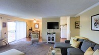 This single-floor space showcases an open-concept layout that seamlessly connects the main living areas. The living room serves as the central hub, featuring plush carpeting, a grey sectional sofa with yellow accent pillows, and a wall-mounted flat-screen television positioned above a white media console with glass-door cabinetry. A textured popcorn ceiling with crown molding detailing runs throughout, adding a classic architectural touch.

Adjacent to the living room, the dining area is finished with tile flooring and accommodates a wooden dining table and chairs beneath a ceiling fan with a light fixture. Vertical blinds on a nearby window allow for adjustable natural light. The area flows naturally into the kitchen, which is visible through an open passageway and features white cabinetry, a white refrigerator, and tiled flooring consistent with the dining space.

The warm beige and yellow wall tones unify the open floor plan, creating a cohesive and inviting atmosphere. The combination of carpeted and tiled flooring zones define each functional area while maintaining an overall sense of continuity throughout the space.