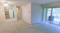 This single-floor residential space offers a clean, neutral interior designed for comfortable everyday living. The main living area is finished with plush wall-to-wall carpeting in a light beige tone, creating a warm and cohesive foundation throughout the space. Smooth, white walls and standard-height ceilings contribute to a bright and airy atmosphere.

A sliding glass door fitted with vertical blinds lines one side of the living area, allowing for natural light and potential outdoor access. A wall-mounted thermostat is conveniently positioned in the main living space, providing easy climate control.

A hallway extends from the living area toward additional rooms, lined with multiple doors that suggest access to bedrooms, closets, or utility spaces. Mirrors are visible along the hallway, adding depth and enhancing the sense of openness. The layout appears functional and straightforward, with a logical flow between spaces.

The overall ambiance of the unit is understated and move-in ready, with a neutral color palette that lends itself well to a variety of furnishing styles. The space is well-maintained, offering a clean canvas suitable for personalization.