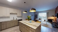 Located on Ferry Road in Warrenville, Illinois, this single-floor residence offers a thoughtfully designed open-concept layout with modern finishes throughout. The kitchen serves as the centerpiece of the home, showcasing a large island with striking veined stone countertops, dual pendant dome lights with warm gold interiors, two-tone cabinetry in light wood and cream tones, a white subway tile backsplash, stainless steel appliances, and ample storage. The kitchen flows seamlessly into the living and dining areas, creating an inviting, connected space bathed in natural light from a large sliding glass door that opens to an outdoor area. The living room features light hardwood-style flooring, neutral walls, and a relaxed, airy ambiance. Two bedrooms offer carpeted flooring, large windows with exterior views, and a calm, restful atmosphere. The primary bedroom is complemented by a spacious walk-in closet with wire shelving and hanging rails for generous storage. The bathroom is finished with large-format tile in the walk-in glass-door shower, wood-toned vanity cabinetry, and hardwood-style flooring, presenting a clean and contemporary aesthetic throughout.