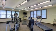 This fitness center offers a practical and well-organized space designed to support a full cardio workout routine. The room is outfitted with multiple treadmills, an elliptical trainer, a stationary bike, and additional cardio equipment, providing users with a variety of exercise options. A wall-mounted flat-screen television is positioned for easy viewing from any machine, enhancing the workout experience. The space is generously lit by a combination of overhead fluorescent strip lighting and natural light streaming in through a row of windows along the perimeter walls, creating a bright and energizing atmosphere. The neutral-toned walls and patterned carpet flooring contribute to a clean, functional aesthetic. The layout is thoughtfully arranged to allow comfortable movement between machines while maximizing the use of available floor space. Overall, this fitness center delivers a focused and practical environment well-suited for daily cardio exercise.
