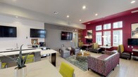 This single-floor amenity space combines a vibrant resident lounge with a fully equipped fitness center, designed with a modern aesthetic highlighted by bold crimson accent walls, light wood-style flooring, and recessed lighting throughout.

The resident lounge is a versatile, open-concept gathering space anchored by a well-appointed kitchen area featuring dark cabinetry, white quartz countertops, a stainless steel refrigerator, oven, and a Starbucks coffee station. A large island with yellow upholstered bar stools provides casual seating, while wall-mounted flat-screen TVs add to the functional design. A warm wood-paneled accent wall and decorative lighting create visual interest. A cozy dining nook, tucked against floor-to-ceiling windows, offers booth-style seating beneath pendant lights and framed artwork, with ample natural light and outdoor views. The main lounge area features plush sectional and club-style seating arranged around a central area rug.

The fitness center boasts soaring ceilings with exposed industrial ductwork and oversized glass garage-style doors that open to views of an outdoor pool area, flooding the space with natural light. Equipment includes ellipticals, cycling bikes, a weight rack with dumbbells and kettlebells, medicine balls, and a multi-function strength training station. A mirrored wall and a mounted TV complete the workout environment.