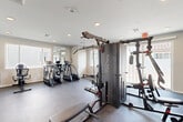 Located at 870 S Sarival Ave in Goodyear, AZ, this dedicated fitness center offers a well-equipped, single-floor exercise space designed to support a comprehensive workout experience. The room features durable rubber flooring throughout, providing a safe and resilient surface ideal for a wide range of physical activities.

Cardio equipment includes multiple treadmills and elliptical trainers positioned near large windows with slatted blinds, allowing ample natural light to fill the space. A rowing machine adds additional cardiovascular options. For strength training, the facility is equipped with a multi-station Body-Solid cable machine, a dedicated weight bench, and a full dumbbell rack with a varied selection of free weights.

Full-length wall mirrors enhance the sense of openness and allow users to monitor their form during workouts. Recessed lighting illuminates the space evenly, complementing the natural light from multiple windows. A wall-mounted television provides entertainment during cardio sessions.

Amenities include a built-in water fountain station with a bottle-fill feature and a trash receptacle nearby. Motivational fitness posters adorn the walls, contributing to an energizing atmosphere. Multiple access doors and clearly marked emergency exits ensure safety and convenience. The clean, neutral color palette and bright interior create a functional, welcoming environment suited for daily fitness use.