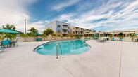 Located at 15350 SW 136th Street in Miami, FL, this outdoor amenity space showcases a expansive, freeform swimming pool as its central feature. The pool's kidney-shaped design accommodates both leisure swimmers and those seeking relaxation, with clearly marked depth indicators and stainless steel entry rails for safe access. Surrounding the pool is a generously sized, light-colored concrete sundeck outfitted with coordinated teal chaise lounge chairs, umbrella-shaded seating clusters, and dining-height patio tables — providing ample space for sunbathing and socializing under the South Florida sky. The deck is enclosed by a white perimeter wall that offers privacy while maintaining an open, airy atmosphere. Tall, manicured royal palm trees are planted at regular intervals along the walkways and within the deck area, lending a distinctly tropical character to the environment. A covered pergola structure is visible along one side of the pool area, providing shaded gathering space. The surrounding multi-story residential buildings feature modern architecture with stone accents, large windows, and private balconies that overlook the pool courtyard. Gated pedestrian access points flanked by palm trees and white stone landscaping complete this well-appointed outdoor retreat.