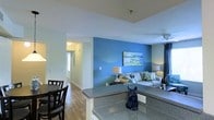 This single-floor residence offers a thoughtfully designed open-concept layout that seamlessly connects the kitchen, dining, and living areas into one cohesive and functional space. The kitchen features a granite-style countertop that doubles as a breakfast bar, providing a natural boundary between the cooking and living zones while maintaining an open, airy feel throughout.

The living area is anchored by a bold blue accent wall that adds a striking pop of color and visual depth to the space. A comfortable sofa with decorative cushions sits beneath a piece of wall art, complemented by coordinating table lamps that create a warm and inviting ambiance. Dark curtain panels frame the windows, adding contrast to the bright, white ceiling and light-toned walls found throughout the rest of the space.

The dining area is positioned adjacent to the kitchen, accommodating a dark wood dining table and chairs that ground the space with a classic, functional aesthetic. Hardwood flooring flows consistently throughout, tying each area together with warmth and continuity.

A hallway visible from the main living area leads toward additional rooms, with natural light filtering in from a door at the end, suggesting easy access to an entry or exterior. The overall ambiance is modern, clean, and welcoming, with a well-balanced blend of bold design choices and neutral finishes.