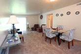 Located at 6200 Hightower Road in Portsmouth, Virginia, this single-floor residence offers a functional and comfortable layout designed to accommodate a variety of living needs. The living room serves as the social heart of the home, featuring plush carpeting, neutral-toned walls, and a large sliding glass door dressed with vertical blinds that floods the space with natural light and provides access to the outdoors, where lush greenery is visible. The galley-style kitchen is efficiently arranged with warm oak cabinetry lining both sides, offering generous storage and counter space. Light tile flooring, a double basin sink, and a full-size refrigerator complete this practical cooking space, which connects seamlessly to adjoining areas. The primary bedroom is generously sized with soft carpeting, a closet, and a window that brings in natural light. A second bedroom accommodates two twin beds comfortably, with plush carpeting, ample floor space, and a large double sliding closet providing substantial storage. The bathroom presents a clean, neutral aesthetic with a window allowing natural light. Throughout the residence, consistent use of neutral wall tones, carpeted floors in living areas, and well-maintained finishes contribute to a cohesive and inviting atmosphere.