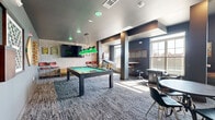 Located at 3700 Monument Avenue in Richmond, Virginia, this entertainment lounge is a well-appointed recreational space designed for social gatherings and leisure. The open-plan layout is anchored by a full-size pool table with signature green pendant lighting, set atop patterned carpet that adds texture and visual interest to the room. Exposed white brick along one wall lends an industrial-chic character, complemented by dark accent columns and a muted gray color palette throughout.

A wall-mounted flat-screen TV provides a focal point for casual viewing, while bar-height seating along the perimeter offers additional spots to relax. Natural light streams in through large windows, brightening the space and offering views of the surrounding neighborhood. A dedicated golf simulator bay, enclosed by heavy curtain dividers and finished with artificial turf flooring, provides an immersive experience for golf enthusiasts. Adjacent to the simulator, a dartboard, compact beverage cabinet, and round table with seating create an intimate gaming nook. The space also features a cozy tufted armchair positioned beside a sun-filled window, ideal for a quiet moment. Together, these elements make this lounge a dynamic, multi-use recreational destination.