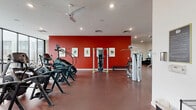 This well-appointed fitness center in Fayetteville, Arkansas offers a comprehensive exercise facility designed to support a wide range of workout routines. The space is defined by a bold design aesthetic, featuring striking red accent walls that energize the environment, complemented by clean white ceilings and durable red rubber flooring throughout. Ceiling fans and recessed lighting ensure the space remains comfortable and well-lit at all times.

The cardio zone is generously equipped with StairMaster climbers, Cybex treadmills, and a row of elliptical machines, providing ample options for aerobic training. A dedicated strength training area houses a variety of Nautilus and free-weight machines, including cable systems, bench press stations, and multi-functional weight machines, allowing for thorough full-body workouts. A functional training zone features a power rack and additional benching options, supported by wall-mounted mirrors that allow users to monitor their form.

Natural light enters through a series of clerestory windows positioned along the upper perimeter of the walls, creating a bright and open atmosphere without compromising wall space used for equipment. A mounted flat-screen television adds a media element to the space. Framed artwork displayed along the red accent wall adds a polished, curated touch to the overall design, elevating the facility beyond a standard gym environment.