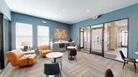 This thoughtfully designed single-floor business lounge and co-working space offers a versatile and professional environment suited for both focused work and collaborative gatherings. The interior is defined by a striking teal-blue color palette accented by warm terracotta and neutral tones, creating a vibrant yet inviting atmosphere throughout.

The open main area features patterned carpet flooring and is furnished with multiple round bistro tables paired with modern chairs, providing flexible seating arrangements for individuals or small groups. A lounge corner with orange leather accent chairs and a side table adds a comfortable, informal touch, while a wall-mounted television and banquette seating in a separate zone offer additional gathering options.

Glass-walled enclosures line one side of the space, housing a private conference room with a large white table, leather rolling chairs, and ample natural light from slatted-blind windows, as well as a dedicated individual workstation pod for focused tasks. These transparent partitions maintain an open, airy feel while providing acoustic separation.

A built-in kitchenette counter runs along one wall, equipped with a coffee maker, microwave, and printer, supporting day-to-day productivity needs. Bold graphic wall art and recessed lighting complete the polished, contemporary aesthetic of this well-equipped communal workspace.