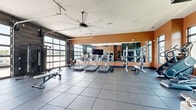 This fitness center offers a comprehensive workout environment across a single floor, thoughtfully divided into two distinct functional zones. The main workout area is a spacious, open room characterized by dark rubber-tile flooring, warm terracotta-toned accent walls, and floor-to-ceiling glass garage-style doors that flood the space with abundant natural light and provide outdoor views. Overhead, a large industrial ceiling fan promotes airflow throughout. This primary zone is outfitted with a row of treadmills, a stair climber, elliptical machines, a cable machine, a full dumbbell rack, weight benches, and a large wall-mounted television. Full-length mirrors along one wall visually expand the space and aid in form correction. A dedicated dumbbell and functional training corner includes a wall-mounted rack stocked with kettlebells, medicine balls, and stability balls, alongside TRX suspension straps. The secondary room features a calmer ambiance with textured wood-panel wall cladding, plush carpeted flooring, and expansive windows overlooking surrounding greenery. This studio space is equipped with stationary cycling bikes and its own wall-mounted display, making it well-suited for focused cycling sessions or group fitness classes. Together, the two zones create a versatile, well-lit, and fully equipped fitness facility.