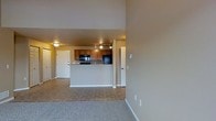 This single-floor space features an open-concept design that seamlessly connects the living area and kitchen. The living room is finished with plush carpeting in a neutral tone, contributing to a warm and inviting atmosphere. Warm beige walls carry consistently throughout the space, creating a cohesive and understated aesthetic.

The kitchen is positioned centrally and defined by a raised peninsula counter that separates it from the living area, offering both a functional workspace and a visual boundary between the two zones. Warm wood-toned cabinetry lines the kitchen, complemented by black appliances including a built-in microwave and refrigerator. A tile floor in the kitchen provides a practical and durable surface that contrasts neatly with the carpeted living area.

Track lighting installed on the kitchen ceiling provides focused illumination, while a flush-mount ceiling fixture offers additional ambient light. Multiple doorways are visible throughout the space, suggesting convenient access to additional rooms. The layout is practical and efficient, making good use of the available footprint while maintaining an open, airy feel.