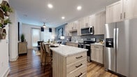 This single-floor residence showcases a contemporary design with consistent wood-look flooring and recessed lighting throughout. The kitchen serves as a central highlight, featuring light wood shaker-style cabinetry with matte black hardware, granite countertops, and a full suite of stainless steel appliances including a side-by-side refrigerator, range, microwave, and dishwasher. A generously sized center island with additional drawer storage offers seating and extra prep space. The kitchen flows seamlessly into a dining area and living room, where large windows with blinds and curtain panels allow natural light to fill the space. The living area features a cool-toned accent wall that adds depth and character to the open layout.

The bedroom is a calm, well-appointed retreat featuring plush carpeting, a large window with ample natural light, and tasteful wall decor. The bathroom is finished with granite-topped cabinetry matching the kitchen aesthetic, wood-look flooring, and a shower-tub combination. A dedicated laundry room houses a full-sized Whirlpool washer and dryer alongside built-in shelving, providing practical storage. The overall ambiance is modern and cohesive, blending neutral tones with contemporary finishes.