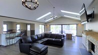 This single-floor space showcases a thoughtfully designed open-concept layout that seamlessly connects the living, dining, and transitional areas. The living area is anchored by a large, dark leather sectional sofa that complements the cool-toned hardwood flooring, creating a grounded yet inviting atmosphere. Overhead, a vaulted ceiling rises dramatically, fitted with multiple skylights that flood the interior with abundant natural light throughout the day. Two wrought-iron candle-style chandeliers hang from the ceiling, adding a rustic yet refined aesthetic to the space. Large sliding glass doors line the far wall, offering unobstructed views of an outdoor deck and the surrounding landscape, blurring the boundary between indoor and outdoor living. Adjacent to the main living area, a dining space is furnished with a wooden table and chairs, positioned to take full advantage of the outdoor views. To the left, a partially visible bar or counter area with seating suggests additional entertaining functionality. The overall ambiance is modern and casual, with a neutral color palette, high-contrast furnishings, and an abundance of natural light contributing to a spacious, airy feel throughout.