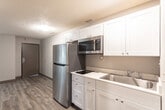 Located at 2530 Washington Blvd in Ogden, Utah, this single-floor unit offers a straightforward, functional layout with a mix of flooring surfaces and neutral color palettes throughout. The living areas feature wood-look plank flooring in a warm gray-brown tone, complemented by light gray walls and white ceilings with a textured finish. Ventilation registers are positioned along the walls and ceilings, indicating a central HVAC system serving the space.

The bedroom areas are finished with a bold patterned carpet in brown and gold tones, providing a distinct contrast to the hard-surface flooring found elsewhere. One bedroom features a window fitted with horizontal blinds, allowing natural light to filter into the room. A separate, smaller carpeted room with textured walls offers additional flexible space.

The bathroom is well-appointed, featuring a soaking tub with a curved front, a full shower curtain rod overhead, a built-in niche for toiletries, and a wood-toned vanity with a white countertop and a mirror above. Wood-look tile flooring ties the bathroom aesthetic to the rest of the unit.

The 3D model reveals a compact but efficiently organized floor plan, with a kitchen area visible toward the rear and clearly defined living and sleeping zones. Overall, the space presents a clean, move-in ready environment with practical finishes throughout.