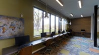 This contemporary computer lab is thoughtfully designed to foster productivity and focus. A long, sleek workstation counter runs the length of the room, positioned directly beneath a continuous bank of large, floor-to-ceiling windows. The generous glazing floods the space with natural light and provides unobstructed views of a lush, tree-lined outdoor area, creating a pleasant and inspiring work environment.

Each workstation is equipped with an all-in-one desktop computer and paired with an ergonomic, cushioned task chair, ensuring both functionality and comfort for users. Overhead, a combination of recessed lighting and distinctive oversized pendant fixtures — shaped like decorative bulbs — adds a modern, stylish accent to the space while providing ample task illumination.

The interior design reflects a vibrant and contemporary aesthetic. Olive green accent walls complement the warm wood tones of the workstation surfaces and cabinetry, while a boldly patterned, multi-toned carpet tile floor adds visual interest and energy to the room. The ceiling integrates linear ventilation ducts and recessed lighting in a clean, organized manner. Overall, the space balances a professional, functional layout with thoughtful design elements that make it both welcoming and highly practical.
