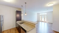 Located at Windsor Station in Windsor, CT, this thoughtfully designed 1-bedroom, 1-bathroom residence offers a functional and modern layout across a single floor. The open-concept kitchen and living area serves as the heart of the home, where rich dark-espresso cabinetry contrasts beautifully with granite countertops that extend across a kitchen island with a built-in sink. Stainless steel appliances, including a refrigerator, range, and microwave, complete the well-equipped kitchen, which is further enhanced by stylish pendant lighting. Warm hardwood-style flooring flows throughout the main living area, leading to a sun-filled living room anchored by a set of glass French doors with transom windows above, opening to a private balcony overlooking lush green surroundings. The bedroom features plush carpeting, a large window with abundant natural light and outdoor views, and direct access to a spacious walk-in closet with built-in wire shelving. The bathroom is finished with matching granite countertops, dark cabinetry, a large mirror with contemporary lighting, and neutral tile flooring, creating a clean and cohesive aesthetic throughout the residence.