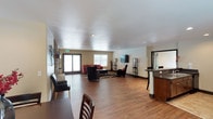 This single-floor space is designed as an open, multi-functional gathering area that balances comfort and practicality. The layout flows seamlessly from a dining area in the foreground — furnished with sturdy wooden chairs — into a spacious lounge zone at the center of the room. The lounge is appointed with a red sofa, cushioned armchairs, and a coffee table arranged on a area rug, creating an inviting seating arrangement anchored by a wall-mounted flat-screen television. Tall, slim shelving units flank the TV, adding both structure and vertical visual interest to the space. The flooring throughout is a warm-toned hardwood, lending a cohesive and polished aesthetic to the entire area. Natural light enters generously through large windows and a sliding glass door along the far wall, brightening the neutral beige and white color palette. To the right, a bar-style counter with dark granite-look surfaces and raised seating provides an additional functional zone, suited for casual interaction or refreshments. The ceilings are smooth and well-lit with recessed lighting, contributing to the clean, modern feel of the interior. Overall, the space presents a well-organized, comfortable environment ideal for communal use.