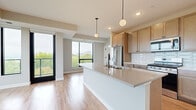 This single-floor residence showcases a contemporary design characterized by clean lines, neutral tones, and an abundance of natural light throughout. The open-concept living and kitchen area serves as the heart of the unit, where large floor-to-ceiling windows frame sweeping views of the surrounding treetops and landscape, flooding the space with daylight. Light wood-tone flooring runs seamlessly through the main living areas, reinforcing the bright, airy atmosphere.

The kitchen is well-appointed with warm wood cabinetry, quartz countertops, a white subway tile backsplash, and a full suite of stainless steel appliances including a gas range, refrigerator, and built-in microwave. A generous island with an integrated double sink provides additional prep space and is illuminated by globe pendant lights overhead.

The bedrooms feature plush carpeting and ceiling fans, with large windows offering continued outdoor views. One additional room features hardwood-style flooring and a corner window configuration, maximizing light exposure. The bathroom is finished with wood-tone cabinetry, matte black hardware, and wood-look flooring. A dedicated laundry room houses a full-size front-loading washer and dryer. Overall, the space presents a polished, modern aesthetic with functional, well-considered finishes throughout.