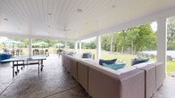 Located at 2000 Day Hill Rd in Windsor, Connecticut, this expansive covered outdoor pavilion offers a well-appointed recreational retreat designed for both relaxation and social activity. The structure features a crisp white shiplap exterior and a matching tongue-and-groove ceiling, giving the space a clean, modern-farmhouse aesthetic throughout.

The stamped concrete flooring spans the entire pavilion, providing a durable and visually appealing foundation that complements the overall design. Open on multiple sides and supported by white structural columns, the pavilion allows for unobstructed sightlines to the surrounding landscape — including a lush green lawn, mature trees, and a waterfront view that enhances the overall ambiance.

The space is organized into distinct functional zones. A generous lounge area features large sectional wicker seating with teal and striped accent cushions, ideal for casual gatherings. Adjacent to this is a dedicated recreation zone with a full-size ping-pong table. A separate dining area with wicker table and chairs is positioned to take advantage of the outdoor views, while additional umbrella-shaded seating is visible on the surrounding grounds.

A built-in white counter with bar-height seating, wall-mounted flat-screen televisions, and ceiling-mounted speakers add modern amenities to the space. Ceiling fans provide comfort throughout, and black-framed French doors provide access to an adjacent interior structure. This pavilion is well-suited for community or recreational use.