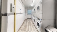 Located at 80 Wellesley Street East in Toronto's Church-Wellesley Village, this shared laundry utility room is a well-maintained, efficiently organized facility designed to serve building residents. The room features a row of stacked, coin-operated washing machines and dryers lining one wall, providing ample laundry capacity. The interior is finished with white subway-style wall tiles accented by a grey horizontal band, creating a clean and functional aesthetic. Light wood-look flooring adds a contemporary touch to the utilitarian space. Overhead fluorescent strip lighting and a flush-mounted ceiling fixture ensure the room is well-lit throughout. A window at the far end of the room allows natural light to filter in, brightening the space. A stainless steel utility sink is conveniently tucked into an alcove, accompanied by a notice board displaying relevant building information and instructions. A prominent yellow gas pipe runs vertically along one wall, and a fire extinguisher is mounted near the entry door for safety. The grey steel entry door is equipped with a window panel. The room is compact yet thoughtfully arranged to maximize usability, making it a practical and accessible amenity for residents of the building.