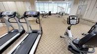 This single-floor fitness center is a comprehensive workout facility designed to support a wide range of exercise needs. The space is divided into distinct functional zones, each thoughtfully arranged to maximize usability. The cardio area is prominently featured near a sweeping wall of floor-to-ceiling windows, which flood the space with natural light and offer views of lush outdoor greenery and an adjacent indoor pool. Treadmills, recumbent bikes, and elliptical trainers are positioned throughout this well-lit section, creating an inviting atmosphere for aerobic exercise. A strength training zone houses a multi-station cable machine, weight benches, and a variety of resistance equipment, providing a complete free-weight and machine workout experience. The flooring transitions between patterned carpet and hard surface in specific zones, and white painted brick columns add architectural character throughout. Wall-mounted mirrors enhance the sense of space and allow users to monitor their form. Recessed panel ceilings with flush lighting keep the interior bright and uniform. Built-in seating ledges along the perimeter offer convenient rest areas. The facility also includes access to locker room doors and utility areas, making it a self-contained and practical fitness destination.