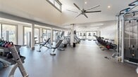 Located at 3680 Corners Mill Dr. in Peachtree Corners, GA, this fitness center offers a comprehensive and thoughtfully designed exercise environment across a single, expansive floor. The space is characterized by soaring ceilings with large clerestory windows that flood the interior with natural light, complemented by a large ceiling fan that promotes air circulation throughout the main workout area. Durable rubber flooring extends throughout, providing both safety and functionality for a wide range of activities. The facility is organized into distinct workout zones. The cardio section features a row of treadmills, ellipticals, and cycling bikes, while a striking black-and-white open-road mural adorns the wall, adding a dynamic visual element to motivate users. A dedicated free weights area includes adjustable benches, a full rack of dumbbells, kettlebells, and medicine balls, along with a wall-length mirror to assist with form. The strength training zone is equipped with multiple cable and resistance machines. Additional functional training space provides room for stretching and floor exercises. Large windows along the exterior walls offer views of the surrounding outdoor area, creating an open and energizing atmosphere throughout the facility.