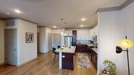 This single-floor residence offers a thoughtfully designed open-concept layout that seamlessly connects the kitchen, dining, and living areas. The kitchen is a standout feature, showcasing a two-tone cabinetry scheme with rich dark wood lower cabinets and crisp white upper cabinets, all complemented by granite countertops with subtle veining. A central island with a built-in sink provides additional workspace and casual seating, while pendant lighting adds a refined touch overhead. Stainless steel appliances, including a refrigerator, microwave, and range, complete the functional yet stylish kitchen. The living area is bathed in natural light through large windows that frame outdoor greenery, creating a warm and inviting atmosphere. Wood-look flooring flows throughout the main living spaces, lending continuity and warmth to the design. The bedroom features plush carpeting, a large sliding glass door with outdoor views, and ample natural light. A dedicated hallway connects to in-unit stacked laundry and a closet with built-in shelving for organized storage. The bathroom is finished with granite vanity countertops, wood-look tile flooring, and a tiled tub-shower combination. The entryway includes a coat closet adjacent to the front door, offering a practical and welcoming arrival experience.