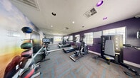 Located at 4000 E Bonanza Rd in Las Vegas, NV, this exercise room is a dedicated fitness space designed to deliver a complete workout experience within a compact footprint. The room features a bold, energizing aesthetic highlighted by deep purple accent walls and a large, full-wall photographic mural depicting a triumphant figure atop a rocky cliff at sunrise — a motivating visual focal point for any workout session. A full-length wall mirror visually expands the space and aids in form correction during exercise. The patterned carpet flooring in blue and tan tones adds a modern, dynamic look underfoot. Cardio equipment is well-represented, with treadmills, an elliptical trainer, and a Freematic spin bike equipped with an interactive display screen. A medicine ball rack with multiple weighted balls provides options for functional strength training, while a wall-mounted yoga/exercise mat offers space for stretching and floor work. Natural light filters in through windows with blinds, offering a pleasant connection to the outdoors. Recessed ceiling lighting ensures the space is well-lit at all times. The room is accessed through a glass-paneled door, and a hand sanitizer dispenser is conveniently mounted near the entrance, emphasizing cleanliness and user well-being.
