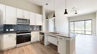 This well-appointed 2-bedroom, 2-bathroom residence offers a thoughtfully designed single-level layout with a clean, contemporary aesthetic throughout. Light wood-style flooring runs consistently across all rooms, creating a cohesive and airy feel.

The open-concept kitchen and living area serves as the heart of the home. The kitchen features white shaker cabinetry, a dark tile backsplash, stainless steel appliances including a refrigerator, range, microwave, and dishwasher, and a spacious peninsula with a neutral-toned countertop and an undermount sink. Three stylish pendant lights hang above the peninsula, adding a modern design touch. A ceiling fan is positioned in the adjacent living area, which benefits from natural light streaming through large, floor-to-ceiling windows framed in black that offer open outdoor views.

The primary bedroom is generously sized and features its own large window with the same bold black framing, allowing abundant natural light. A dedicated walk-in closet with built-in shelving and hanging rods provides ample storage. The two bathrooms are finished with sleek, modern vanities featuring undermount sinks and neutral countertops. The layout also includes a dedicated closet for additional storage needs.