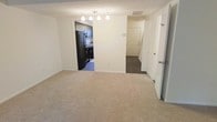 This two-floor residence offers a functional and well-organized layout suited for everyday living. The main living area features neutral wall-to-wall carpeting that adds warmth underfoot, complemented by clean white walls and a textured ceiling. Pendant lighting hangs above, providing focused illumination and a subtle decorative touch to the space.

Adjacent to the living area, the kitchen is accessible through an open doorway and is outfitted with full-size black appliances, including a refrigerator and range, set against dark cabinetry. The kitchen floor transitions to a hard surface, offering a practical contrast to the carpeted living space.

Near the entryway, a paneled front door provides a welcoming entry point, with a staircase visible nearby that connects to the second floor. A closet with louvered doors adds convenient storage directly off the entry area. The overall color palette is neutral and understated, with white trim and doors throughout contributing to a clean, cohesive aesthetic.

The layout is straightforward and efficient, making excellent use of the available space across both floors. The open sightlines between the living room and kitchen promote a connected feel, while distinct zones for entry, storage, and daily living ensure the home remains practical and easy to navigate.