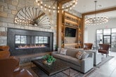 Located at 303 Arrowhead Lane in South Setauket, NY, this sophisticated single-floor clubhouse offers a richly appointed gathering space designed for comfort and community. The main lounge commands attention with soaring ceilings, a dramatic stone accent wall framing a linear fireplace, and a striking multi-bulb chandelier that casts warm ambient light across the room. Floor-to-ceiling built-in shelving with integrated LED lighting flanks a large mounted television, while leather and upholstered seating arrangements invite relaxed conversation.

A secondary lounge area features bold teal shiplap paneling adorned with curated portrait artwork and additional media display, creating an intimate yet stylish retreat. Transitioning toward the event and dining spaces, the clubhouse opens into a generously sized room defined by expansive black-framed floor-to-ceiling windows that flood the interiors with natural light and frame views of the surrounding community. A long communal table with navy upholstered chairs anchors the space beneath an elegant chandelier, while smaller seating clusters and a cozy bistro nook with round tables offer flexible gathering options.

The kitchenette area features sleek cabinetry, a sage-toned countertop with a gold undermount sink, and a textured tile backsplash. Throughout the clubhouse, warm hardwood and stone tile flooring, high ceilings, and carefully curated furnishings contribute to an overall ambiance that is both refined and welcoming.