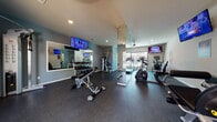 This single-floor fitness center is thoughtfully designed to support a full range of workout routines. The space is divided into distinct functional zones, each tailored to a specific type of exercise. The cardio area is equipped with multiple Cybex treadmills and elliptical machines, positioned alongside large windows that invite natural light and provide views of the outdoor surroundings. A ceiling fan overhead keeps the area comfortable during intense sessions, and a wall-mounted TV ensures entertainment while exercising.

The strength training zone features a multi-functional cable machine, an adjustable workout bench, and a well-stocked dumbbell rack with a range of weights. A large wall mirror visually expands the space and allows users to monitor their form. An additional TV is mounted in this area as well, maintaining a consistent, engaging atmosphere throughout the facility.

Medicine balls and stability equipment are neatly organized along one wall, offering versatility for functional training. The walls are painted in calming blue-gray tones, complemented by recessed and ceiling lighting that keeps the space bright and welcoming. Durable rubber flooring covers the entire facility, providing safety and impact absorption. Double doors at one end open to the outdoors, enhancing ventilation and connectivity with the exterior. A motivational fitness-themed graphic adorns one wall, reinforcing the energetic character of the space.