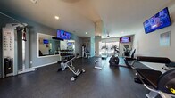 This single-floor fitness center is thoughtfully designed to support a full range of workout routines. The space is divided into distinct functional zones, each tailored to a specific type of exercise. The cardio area is equipped with multiple Cybex treadmills and elliptical machines, positioned alongside large windows that invite natural light and provide views of the outdoor surroundings. A ceiling fan overhead keeps the area comfortable during intense sessions, and a wall-mounted TV ensures entertainment while exercising.

The strength training zone features a multi-functional cable machine, an adjustable workout bench, and a well-stocked dumbbell rack with a range of weights. A large wall mirror visually expands the space and allows users to monitor their form. An additional TV is mounted in this area as well, maintaining a consistent, engaging atmosphere throughout the facility.

Medicine balls and stability equipment are neatly organized along one wall, offering versatility for functional training. The walls are painted in calming blue-gray tones, complemented by recessed and ceiling lighting that keeps the space bright and welcoming. Durable rubber flooring covers the entire facility, providing safety and impact absorption. Double doors at one end open to the outdoors, enhancing ventilation and connectivity with the exterior. A motivational fitness-themed graphic adorns one wall, reinforcing the energetic character of the space.