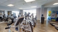 This single-floor fitness center offers a comprehensive workout environment designed to accommodate a variety of exercise routines. The space is generously filled with modern cardio equipment, including elliptical trainers, stationary bikes, and additional strength-training machines arranged in an orderly layout that allows for easy movement throughout the facility.

Large, floor-to-ceiling windows line the walls, flooding the interior with abundant natural light and offering open views of the surrounding outdoor landscape. The bright, airy atmosphere is further enhanced by a drop-panel ceiling with recessed lighting, ensuring the space remains well-lit at all hours.

The flooring consists of warm hardwood-style planks, providing a clean and durable surface suited to high-traffic fitness use. The walls are finished in a neutral light gray tone, contributing to a calm and focused workout environment. A small amenity station is positioned near the perimeter, offering convenience for users during their sessions.

Overall, the fitness center presents a modern, welcoming, and functional space that balances practicality with a bright, open ambiance — making it an inviting destination for daily exercise.