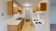 This single-floor unit offers a straightforward, functional layout designed for comfortable everyday living. The living and bedroom areas are finished with wall-to-wall carpet in a neutral tone, creating a warm and cohesive feel throughout. Baseboard heating runs along the exterior walls, ensuring efficient climate control, while a window-mounted air conditioning unit provides cooling during warmer months. Ceiling fans with light fixtures are present in the bedroom areas, adding both airflow and ambient lighting. Windows allow natural light to filter into the rooms, keeping the spaces feeling open despite their modest scale.

The galley-style kitchen is well-appointed with warm oak cabinetry lining both walls, offering generous upper and lower storage. White appliances — including a coil-burner electric range, refrigerator, and range hood — are neatly integrated alongside a double-basin sink with a clean, light-colored countertop. The kitchen connects to an interior area with additional closet storage. The bathroom features a full-sized soaking tub with an overhead shower and curtain rod, tile flooring, and standard fixtures in a clean white finish. The overall aesthetic is simple, neutral, and move-in ready.