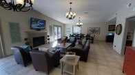 This single-story space features an open-concept layout that seamlessly connects the living and dining areas, creating a generous, flowing environment ideal for both everyday living and entertaining. The living area is anchored by a brick fireplace and includes comfortable dark leather seating arranged around a central coffee table, with a wall-mounted flat-screen television above the mantle. Vaulted ceilings add a sense of height and openness, while a textured ceiling treatment adds subtle architectural character throughout. Two elegant chandelier-style light fixtures illuminate both the living and dining zones, enhancing the warm, welcoming ambiance. The dining area accommodates a full-sized table with seating for multiple guests, positioned beneath its own chandelier. Light tile flooring runs throughout, contributing to a bright and clean aesthetic. French doors along the back wall invite natural light into the space and provide direct access to an outdoor area, further extending the sense of openness. Decorative wall art and tasteful accents add personality without overwhelming the overall cohesive, neutral color palette.