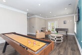 This inviting single-floor recreational space is thoughtfully designed for both entertainment and relaxation. The room is defined by rich, dark hardwood flooring that runs throughout, complemented by crisp white crown molding and clean baseboards that frame the space with a polished finish. A striking teal accent wall serves as a bold focal point, anchoring the lounge area where a wall-mounted flat-screen TV is positioned for easy viewing. Recessed lighting in the ceiling provides warm, even illumination across the entire room.

The space is well-equipped for recreation, featuring a full-length shuffleboard table positioned near a bank of windows that flood the room with natural light, and a foosball table situated near glass French doors that open to an outdoor area with lush greenery visible beyond. A casual seating arrangement of golden-yellow leather chairs surrounds a round decorative coffee table, creating a comfortable gathering spot at the heart of the room.

A separate nook area, finished in the same neutral tones, offers a rustic round high-top table with industrial-style adjustable stools — ideal for casual conversation or tabletop games. Multiple windows fitted with white horizontal blinds ensure abundant natural light throughout, while the overall design strikes a balanced blend of modern amenities and warm, welcoming character.