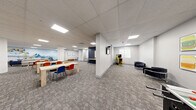Located at 220 Ira Needles Blvd in Kitchener, Ontario, this generously sized single-floor space offers a versatile, open-concept layout designed to accommodate a variety of activities and social interactions. The interior is finished with neutral grey carpeting throughout most of the floor, complemented by tile flooring in the kitchenette area, and a suspended ceiling with recessed panel lighting that ensures consistent, bright illumination across the entire space.

The layout is divided into distinct functional zones. A lounge area features grey sofas and blue accent chairs arranged around a coffee table, creating a comfortable gathering spot. Adjacent to this is a dining and activity area with light wood tables paired with vibrant multi-coloured chairs, ideal for group use. A well-appointed kitchenette includes white cabinetry and a stainless steel refrigerator, supporting everyday convenience.

Architectural columns are integrated throughout the open floor plan, adding structural character while subtly defining zones. Accent walls in deep navy blue provide bold contrast against the light grey walls, and colourful artwork and murals are displayed throughout, lending the space a warm, engaging atmosphere. A quieter retreat area features low-profile seating and curated artwork for a more relaxed setting. A classic arcade game and a piano add recreational and cultural elements. A staircase with metal railings connects to adjacent levels.