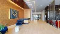 This refined, single-floor venue makes an immediate impression with its grand entrance lobby, where light hardwood flooring, geometric LED ceiling details, and a dramatic copper-toned accent wall set an upscale tone. A sleek reception desk clad in dark marble anchors the space, accompanied by curated seating including bold blue velvet chairs and marble accent tables. Floor-to-ceiling glass entry doors invite abundant natural light and offer clear sightlines to the surrounding streetscape.

Moving through the space, the bar area commands attention with a long counter lined by stylish leather-and-wood bar stools framed by an elegant shelving installation of metal and glass, accented with brass details and pendant lighting. Decorative geometric patterned tile flooring and dark marble surfaces reinforce the space's luxurious character, while chain-link privacy screens add a layer of architectural drama.

The expansive lounge and dining area features large-format polished tile flooring, oversized windows delivering sweeping street views, and a mix of intimate seating arrangements alongside a central communal table. Lush potted plants punctuate the interior throughout, softening the contemporary aesthetic. The overall ambiance is polished and welcoming, balancing modern design with warm, inviting details.