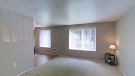 This single-story space presents a clean, functional layout with a welcoming, neutral aesthetic. The main living area is defined by soft, wall-to-wall carpeting and warm gray walls that create a calm, understated ambiance. Two large windows dressed with vertical blinds line the exterior wall, allowing generous amounts of natural light to fill the room while offering views of outdoor greenery. A ceiling fan is positioned overhead, adding practical comfort to the space.

A textured popcorn ceiling adds a classic residential character throughout. The transition between the carpeted living area and an adjacent zone with hardwood-style flooring is visible, suggesting a natural separation between living and dining or entryway spaces. White baseboards provide a crisp contrast against the gray walls and flooring.

Wall-mounted utility fixtures, including a small vent and thermostat panel, indicate practical climate control features integrated seamlessly into the design. The overall atmosphere is bright, airy, and move-in ready, well-suited for a variety of furnishing styles. The space balances simplicity with functionality, offering a versatile foundation for personalization.